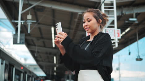 Una-Mujer-Con-Un-Top-Negro-Con-Hombros-Descubiertos-Espera-En-El-Andén-De-Un-Tren,-Absorta-En-Su-Teléfono-Inteligente,-Acercándose-Al-Tren-En-El-Fondo,-Añade-Un-Elemento-Dinámico-Al-Entorno-Urbano.