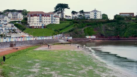 Scenic-Babbacombe-view-of-people-walking-on-the-beach-and-ocean-with-colorful-seaside-huts-and-houses-on-hill-in-Devon,-England-UK