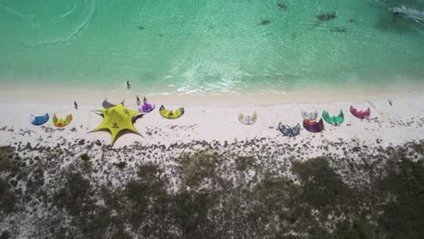 Colorful-kites-and-campers-on-a-white-sandy-beach-with-turquoise-water,-aerial-view