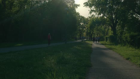 People-walking-along-a-shaded-path-in-the-late-afternoon-at-Jarun-Lake-in-Zagreb,-Croatia,-surrounded-by-lush-greenery