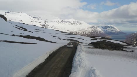 Toma-Aérea-De-Seguimiento-De-Un-Automóvil-Blanco-Conduciendo-Por-Una-Carretera-De-Montaña-Durante-Un-Día-Nevado-De-Primavera-En-Islandia.