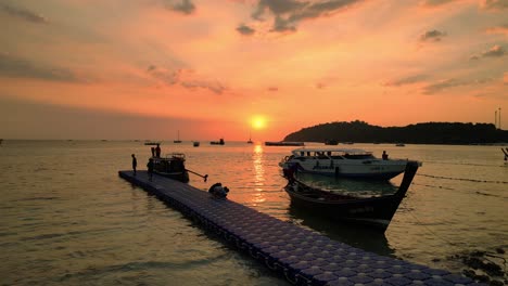 The-sun-sets-over-the-calm-ocean,-casting-a-warm-glow-on-boats-and-an-island-in-the-distance,-painting-the-sky-in-shades-of-orange-and-pink