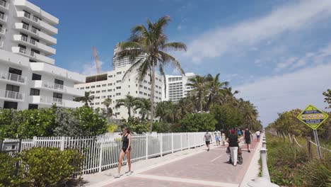 Sonniger-Tag-Am-Miami-Beach-Mit-Menschen,-Die-Entlang-Des-Strandwegs,-Gesäumt-Von-Modernen-Weißen-Gebäuden-Und-Palmen,-Spazieren-Oder-Rad-Fahren