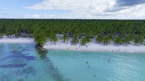 Eine-Drohne-Nähert-Sich-Einem-Strand,-Der-Mit-Weißen-Stühlen-Gesäumt-Ist,-An-Denen-Sich-Touristen-Versammeln,-Während-Einige-Im-Kristallklaren-Blauen-Wasser-Der-Karibik-In-Der-Dominikanischen-Republik-Schwimmen