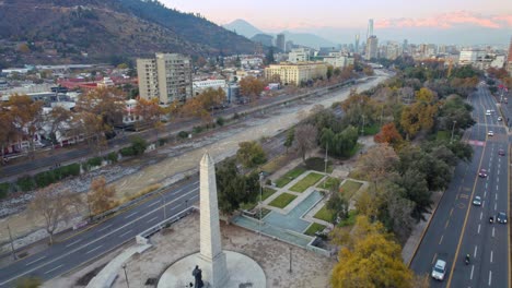 Aerial-Drone-fly-Providencia-Obelisk,-traffic-in-Santaigo-de-Chile,-Andean-City-Avenue-traffic-below-mountain