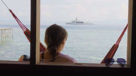 Woman-relaxes-on-Kri-Island-in-the-Raja-Ampat-archipelago,-Indonesia,-gazing-at-a-distant-luxurious-yacht