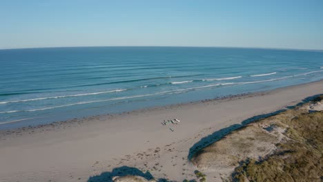 Surfer-Tragen-Bretter-An-Einem-Ruhigen-Strand-Entlang,-In-Der-Ferne-Brechen-Die-Wellen,-Luftaufnahme