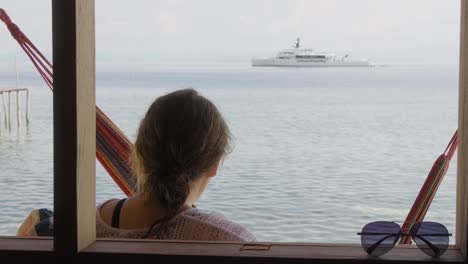 A-woman-relaxes-on-Kri-Island-in-Raja-Ampat-archipelago,-Indonesia,-gazing-out-at-a-tranquil-sea-with-a-yacht-in-the-distance