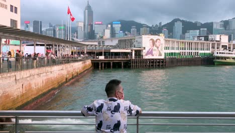 Un-Hombre-Se-Reclina-Sobre-Una-Valla-En-El-Puerto-Y-El-Muelle-De-Victoria,-Disfrutando-Del-Pintoresco-Paisaje-Urbano-Como-Telón-De-Fondo.