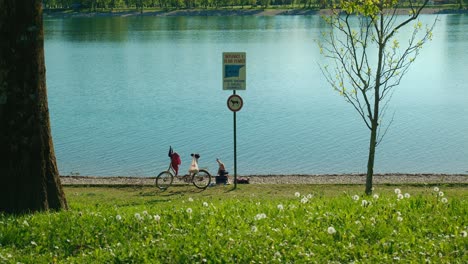 People-resting-by-the-shoreline-of-Jarun-Lake-in-Zagreb-with-bicycles,-grassy-area,-and-a-signpost-in-the-foreground