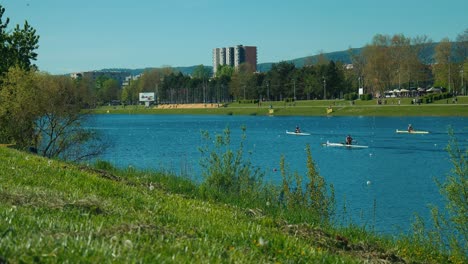 Rowers-on-Jarun-Lake-in-Zagreb,-with-grassy-banks,-trees,-and-city-buildings-in-the-background-on-a-sunny-day