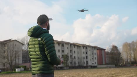 El-Piloto-Del-Dron-Mira-Hacia-Arriba-Mientras-El-Dron-Desciende-Para-Aterrizar-En-Un-Campo-Cerca-De-Los-Apartamentos.