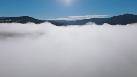 White-layer-of-fog-coats-forest-with-reflection-of-sun-off-river-in-the-Pacific-Northwest-mountains,-Washington-State,-aerial-drone-shot