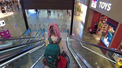 Traveler-with-pink-hair-descends-escalator-at-Istanbul-Airport,-shops-and-people-in-background