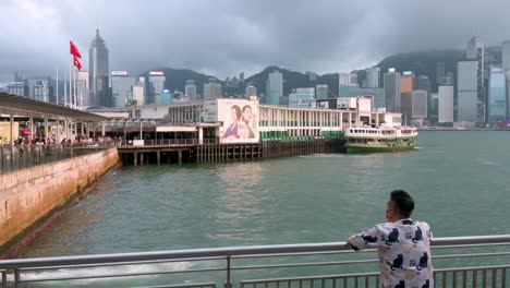 At-Victoria-Harbour-and-pier,-a-man-reclines-on-a-fence,-enjoying-the-view-with-the-cityscape-as-a-backdrop