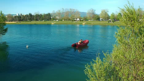 Persona-Remando-Un-Bote-Rojo-En-Las-Tranquilas-Aguas-Del-Lago-Jarun-En-Zagreb,-Rodeado-De-Exuberante-Vegetación-Y-Montañas-Distantes