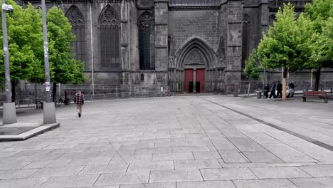 Pedestrian-crossing-a-square-with-side-view-over-black-cathedral-with-red-door-and-green-trees