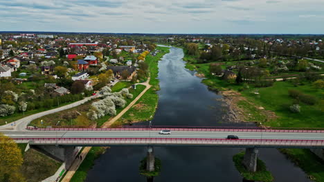 Aerial-drone-pan-shot-over-cars-passing-by-a-bridge-on-Musa-river-alongside-a-town-in-Latvia-on-a-cloudy-day