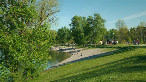 People-relaxing-on-a-sunny-beach-at-Jarun-Lake-in-Zagreb,-Croatia,-surrounded-by-lush-green-trees-and-grassy-areas