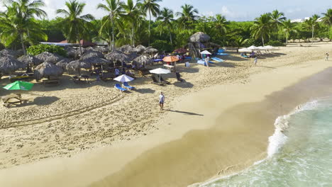 Retreating-and-pulling-up-pedestal-drone-shot-of-a-beachfront-in-Dominican-Republic,-showing-some-tourists-strolling-along-the-beach-and-the-lush,-green-vegetation-in-the-background