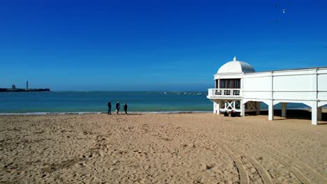 Aerial-view-of-the-beach-in-Cadiz-with-3-people-standing-near-the-water-and-seagulls-flying-all-around