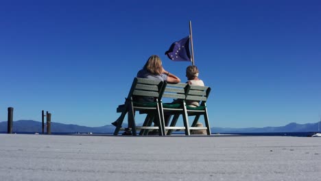 Tiefaufnahme-Von-2-Frauen,-Die-Am-Ende-Des-Docks-Sitzen-Und-Die-Flagge-Alaskas-Wehen