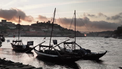 handheld-view-of-three-rabelo-sail-boats-moored-at-Douro-river,-at-dusk,-with-cityscape-and-Arrabida-bridge