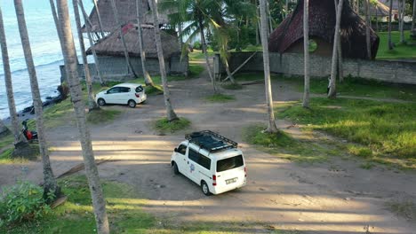 white-camper-van-parked-in-a-coconut-tree-field-overlooking-a-beach-in-Bali,-aerial