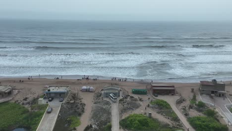 Drohne-Fliegt-über-Das-WWF-Feuchtgebietszentrum-In-Karatschi---Häuser-Am-Strand-Und-Man-Sieht-Das-Meer-Mit-Großen-Wellen-Und-Menschen,-Die-Am-Strand-Stehen