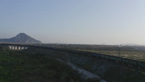 Full-loaded-coal-cargo-freight-train-locomotive-stationary-along-a-high-rise-railway,-in-green-fields-with-mountain-ridge-in-the-far-distance,-on-the-sunset