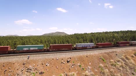 Aerial-cargo-trail-races-by-a-man-standing-by-the-parallel-railroad-tracks-near​-Williams,-Arizona