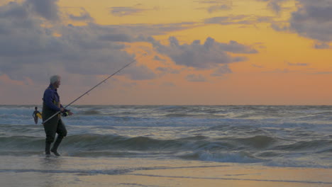 Old-man-is-fishing-on-the-beach-throwing-bait-with-fishing-rod-in-the-surf-during-sunset