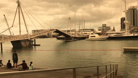 Un-Cielo-Nublado-Proyecta-Un-Tono-Naranja-Sobre-La-Costa-De-Auckland-Mientras-El-Puente-Basculante-Se-Abre-Hacia-Arriba-Mientras-Algunos-Turistas-Observan