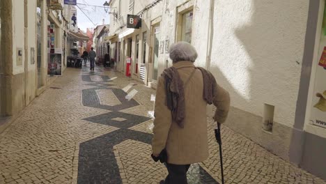 ältere-Frau-Zu-Fuß-Auf-Bürgersteig-Mit-Einem-Stock-In-Einer-Gasse-In-Faro,-Portugal