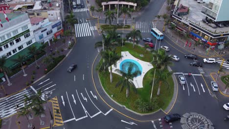 Drone-hovers-above-a-round-about-near-a-public-park-called-"Parque-kennedy"-Camera-tilts-up-showing-the-park-filled-with-many-trees-in-between-two-streets