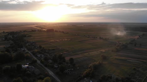 Vista-Aérea-De-Un-Pequeño-Pueblo-Agrícola-Con-Humo-De-Un-Incendio-Durante-La-Puesta-De-Sol