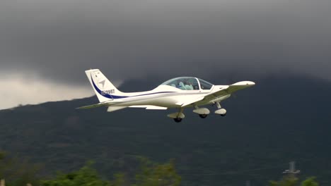 White-two-seat-airplane-taking-off-from-small-air-strip-flying-towards-mountains-on-cloudy-day