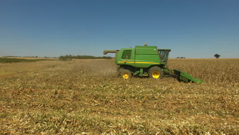 Wide-aerial-shot-at-eye-level-of-the-side-of-a-John-Deere-combine-harvester-tractor-with-spinning-corn-heads-moving-across-a-golden-field-that-rotates-around-the-tractor-to-show-the-front