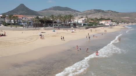 Summertime-scene-of-people-swimming,-relaxing-and-enjoying-ocean-sea-waves-on-Porto-Santo-island-sandy-beach-in-Madeiran-archipelago-with-mountain-range-in-background,-Portugal,-handheld-pan-right