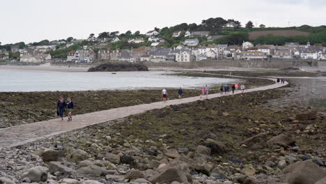 Tourists-walking-on-the-causeway-that-leads-to-Marazion-from-the-english-medioeval-castle-and-church-of-St-Michael's-Mount-in-Cornwall-on-a-cloudy-spring-day,-Zoom-in-4k-footage