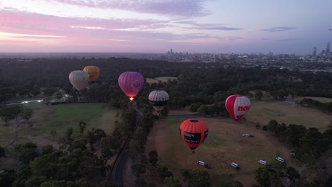 Drone-footage-of-hot-air-balloons-taking-off-at-sunrise-over-green-parklands-in-Melbourne,-Australia