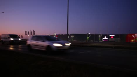 Vehículos-De-Transporte-Que-Viajan-Por-La-Carretera-Asfaltada-En-Wroclaw,-Polonia-Por-La-Noche-Con-Edificios-Comerciales-Al-Fondo---Toma-Amplia