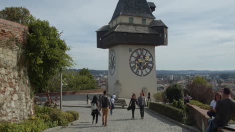 Tilting-shot-of-Schlossberg-clock-tower-with-tourists-passing-by-and-sitting-on-the-walls