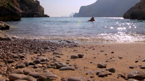 Niedrige-Winkelansicht-Der-Wellen,-Die-Am-Kieselstrand-Brechen,-Mit-Menschen,-Die-Im-Hintergrund-Schwimmen