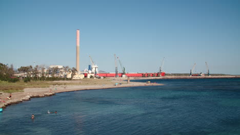 Beach-full-of-people-with-industry-in-background-in-Portovesme,-Portoscuso,-Sardinia,-static,-day