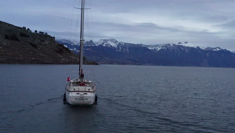 Drone-Siguiendo-Un-Velero-En-Un-Lago-Tranquilo-Con-Montañas-Cubiertas-De-Nieve-Al-Fondo