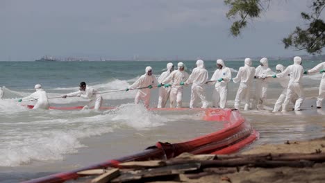 Oil-Spill-At-Mae-Ram-Phueng-Beach,-Navy-Officers-In-PPE-Suit-Pulling-The-Oil-Boom-Barrier-Against-The-Waves-To-The-Beach-In-Rayong,-Thailand