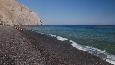 Toma-Amplia-De-Mano-De-La-Playa-De-Perissa-Con-Arena-De-Lava-Negra-Con-Turistas-Tomando-El-Sol,-Nadando-Y-Divirtiéndose-Con-Una-Montaña-Al-Fondo-Y-Cabañas-Tiki-Con-Sombra-Para-El-Sol