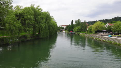 Drone-flying-over-Ljubljanica-river-with-trees-on-both-sides-with-small-waves-on-water-and-people-walking-by-the-river