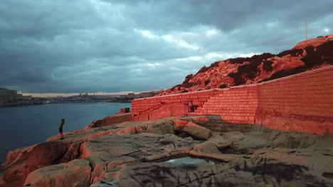 Hombre-Estirándose-Y-Disfrutando-De-La-Hermosa-Vista-Al-Mar-En-Un-Mágico-Amanecer-Rojo-En-Un-Acantilado-En-Malta